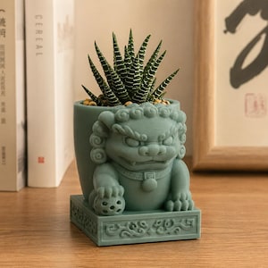 May include: A matte sage green planter shaped like a guardian lion, or shishi, resting on a rectangular base with intricate relief carvings. The planter holds a spiky, dark green succulent with horizontal white stripes on its pointed leaves.