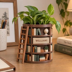 May include: A brown cylindrical planter designed to look like a multi-tiered bookshelf, featuring miniature books, a small globe, a tiny potted plant, and a ladder leaning against the side. The center shelf displays a rectangular sign reading WILLIAMS.