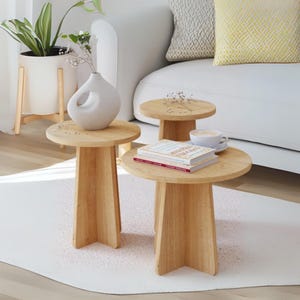 May include: Three light-colored wooden side tables of varying heights. The tables have round tops and flared, geometric legs. One table holds a white vase with a unique cutout design. Another table has books and a cup of coffee. The tables are on a white rug.