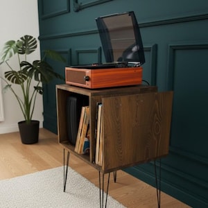 May include: A wooden record player with a clear cover sits atop a matching wooden cabinet. The cabinet has a door and contains vinyl records. The cabinet is supported by black hairpin legs. A potted plant is to the left.