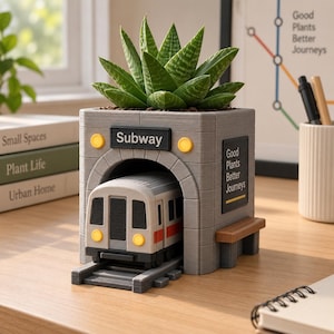 May include: A gray planter shaped like a subway tunnel entrance with a green succulent planted on top. The structure features a miniature train emerging from the tunnel, yellow lights, and signs that read Subway and Good Plants Better Journeys.