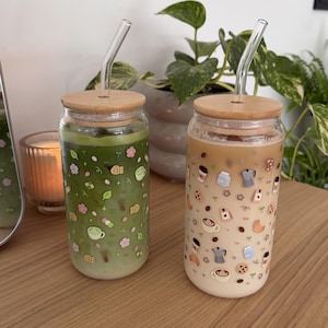 Könnte beinhalten: Zwei klare Glastrinkbecher mit Bambusdeckeln und gebogenen Glasstrohhalmen. Ein Becher enthält ein grünes Matcha-Getränk mit kleinen aufgedruckten Symbolen von Matcha-Schalen, Blättern und Blüten. Der andere enthält ein beiges Kaffeegetränk mit Symbolen von Kaffeetassen, Milchkartons und Keksen.