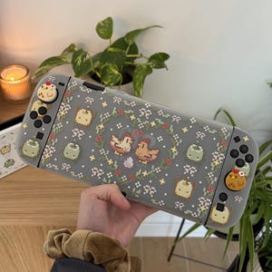 Pode incluir: Uma capa protetora cinza para console de jogos com um padrão de pixel art de galinhas, ovos e elementos florais. A capa inclui protetores de joystick correspondentes com designs de galinhas sobre o dispositivo portátil.