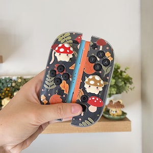 Può includere: Una coppia di controller joy-con racchiusi in cover protettive trasparenti con sfondo scuro, caratterizzati da un motivo di foglie autunnali e piccoli funghi, arricchiti da due ciondoli a forma di fungo in rilievo 3D rossi e marroni.