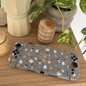 May include: A grey gaming console featuring a repeating pattern of coffee cups, milk cartons, croissants, and cookies. The console has black buttons and joysticks, with one joystick featuring a blue accent and the other featuring a red accent.
