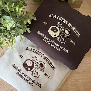 Op de afbeelding: Een donkerbruin sweatshirt en een lichtbeige sweatshirt, beide voorzien van geborduurde tekst met BLATHERS MUSEUM EST 2002 en Exhibition of insects, fish, fossils and art, vergezeld door kleine grafische iconen van een vlinder, bot en vis.