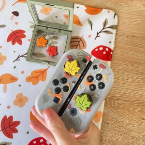 May include: A light gray gaming controller shell featuring a printed autumn leaf and mushroom pattern, equipped with two yellow and green leaf-shaped thumbstick covers, alongside a small olive green display case containing two orange leaf-shaped charms.