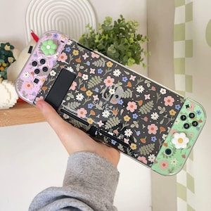 Peut inclure: Une console Nintendo Switch dotée d'une coque de protection noire avec un motif floral coloré composé de fleurs roses, jaunes et blanches. La manette gauche est violet clair et la droite est vert clair, toutes deux avec des capuchons de joystick en forme de fleur blanche.