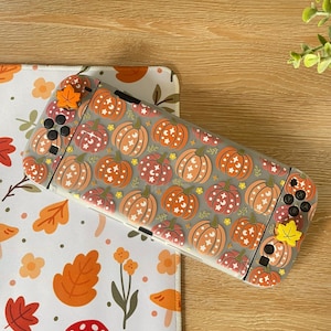 May include: A handheld gaming console featuring a translucent protective case decorated with a repeating pattern of orange and red pumpkins, small white stars, and crescent moons, accented by small orange and yellow leaf-shaped joystick caps.