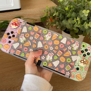 Puede incluir: Una consola de juegos portátil con una funda protectora decorada con un patrón repetitivo de iconos de temática otoñal que incluyen calabazas, fantasmas, hojas, calcetines y tazas de café, con cubiertas de joystick en forma de seta.