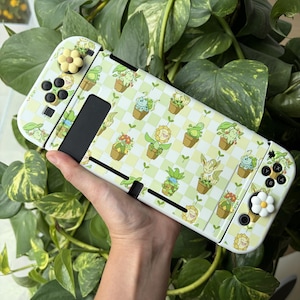 Op de afbeelding: Een draagbare spelcomputer met een lichtgroene en witte geblokte skin met diverse illustraties van planten in potten. Het apparaat bevat twee witte bloemvormige joystickcovers en zwarte knoppen voor een decoratief uiterlijk.