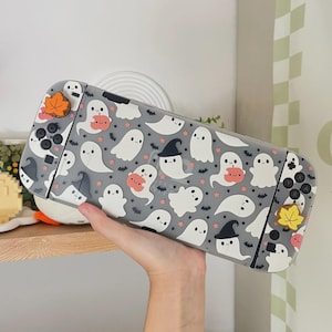 May include: A grey gaming console protective case featuring a repeating pattern of white ghosts, some wearing black witch hats, interspersed with small orange pumpkins, tiny black bats, and orange stars. The case includes matching thumb grips.
