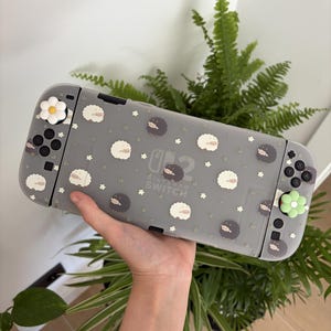 Può includere: Una custodia per console Nintendo Switch grigia traslucida con un motivo ripetuto di piccole pecore bianche e grigio scuro, minuscoli fili d'erba verdi e piccoli fiori bianchi. I copri-levette del joystick hanno la forma di fiori.