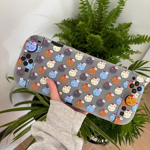May include: A handheld gaming console featuring a translucent gray protective case decorated with a repeating pattern of small, colorful cartoon chickens. The device includes matching joystick covers with chicken designs on both the left and right controllers.