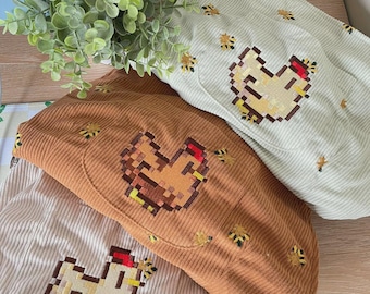 Può includere: Tre pochette in velluto a coste nei colori verde chiaro, marrone e crema, ciascuna con un motivo a pixel ricamato raffigurante un pollo e piccole api. Il tessuto presenta una trama a coste verticali e ogni pochette ha una cerniera.