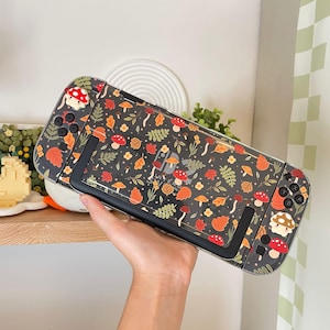 May include: A handheld gaming console featuring a clear protective case with a dark background pattern of colorful autumn leaves, small orange and red mushrooms, and tiny yellow flowers. The design includes matching mushroom-shaped joystick caps.