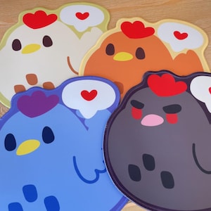 May include: Four mouse pads shaped like cartoon chickens in white, orange, blue, and dark grey. Each chicken features a red comb, a yellow beak, and a white speech bubble containing a small red heart. The designs are flat with smooth edges.