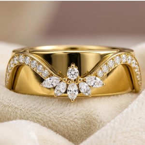 May include: A gold band ring featuring a curved, elevated center section adorned with a cluster of marquise-cut and round-cut clear stones. The band displays a polished finish with additional round-cut stones set along the sweeping, arched edges.