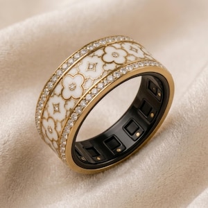 May include: A wide gold smart ring featuring a white enamel band with a repeating four-leaf clover pattern. The ring is bordered by two rows of small clear crystals and has a black interior lining with visible electronic sensors and charging points.