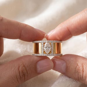 Può includere: Un anello in oro e diamanti con un diamante centrale a taglio marquise. L'anello ha una fascia larga con barre verticali in oro ed è incrostato di piccoli diamanti. L'anello è tenuto tra due dita.