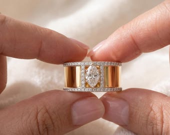 May include: A gold and diamond ring featuring a marquise-cut diamond center stone. The ring has a wide band with vertical gold bars and is encrusted with small diamonds. The ring is held between two fingers.