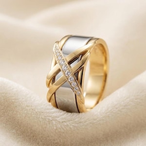 May include: A two-tone ring featuring a wide silver-colored band overlaid with a polished gold-colored crisscross design. One of the diagonal gold-colored bands is accented with a row of small, clear, sparkling stones set along its length.