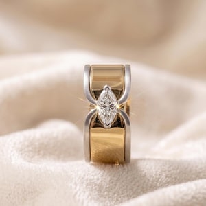 Peut inclure: Une bague bicolore avec une pierre centrale en diamant taille marquise. La bague présente un anneau en or avec des accents argentés et un diamant étincelant. La bague est posée sur un fond doux et neutre.