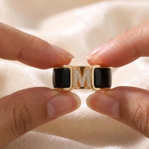 Puede incluir: Un anillo dorado con una banda de esmalte negro y la letra "M" incrustada con pequeños cristales transparentes. El anillo se sostiene entre dos manos, mostrando su diseño. El diseño del anillo es moderno y elegante.