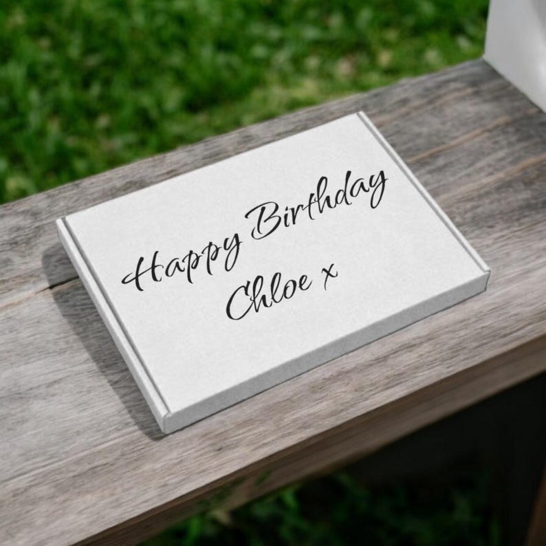 Puede incluir: Una caja de regalo blanca con las palabras "Happy Birthday Chloe x" en escritura negra. La caja est&aacute; sobre una superficie de madera envejecida, con un fondo verde borroso.
