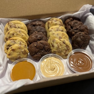 Puede incluir: Una caja de galletas variadas, incluyendo con chispas de chocolate y doble chocolate, dispuestas con tres salsas peque&ntilde;as para mojar. Las galletas son de color marr&oacute;n dorado y marr&oacute;n oscuro, con chispas de chocolate visibles. Las salsas son de caramelo, mantequilla de cacahuete y chocolate.