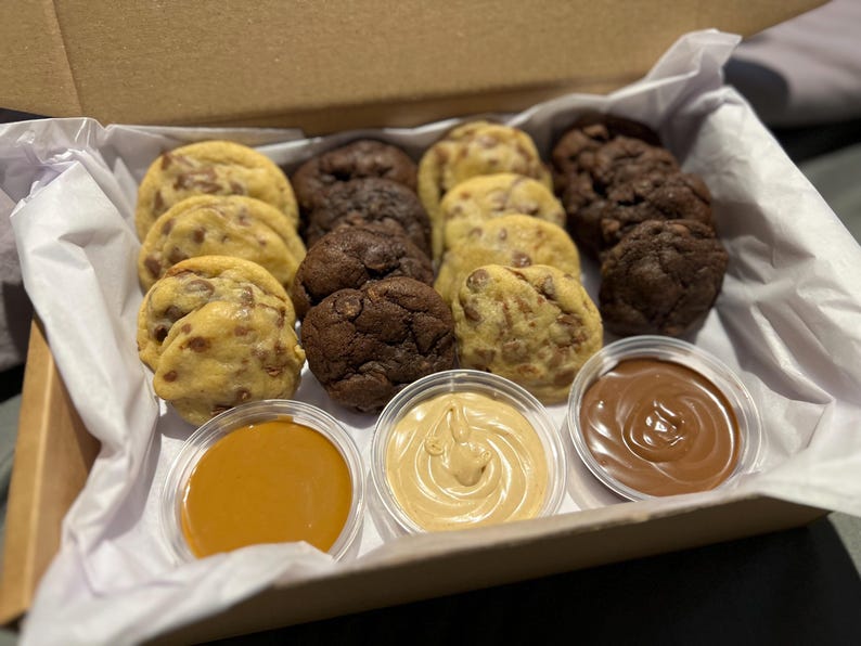 Puede incluir: Una caja de galletas variadas, incluyendo con chispas de chocolate y doble chocolate, dispuestas con tres salsas peque&ntilde;as para mojar. Las galletas son de color marr&oacute;n dorado y marr&oacute;n oscuro, con chispas de chocolate visibles. Las salsas son de caramelo, mantequilla de cacahuete y chocolate.