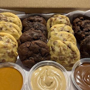 Puede incluir: Una caja de galletas variadas, incluyendo con chispas de chocolate y doble chocolate, con peque&ntilde;os recipientes de salsas para mojar. Las galletas son de color marr&oacute;n dorado y marr&oacute;n oscuro, con chispas de chocolate visibles. Las salsas son caramelo, mantequilla de cacahuete y chocolate.