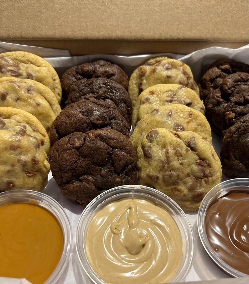 Puede incluir: Una caja de galletas variadas, incluyendo con chispas de chocolate y doble chocolate, con peque&ntilde;os recipientes de salsas para mojar. Las galletas son de color marr&oacute;n dorado y marr&oacute;n oscuro, con chispas de chocolate visibles. Las salsas son caramelo, mantequilla de cacahuete y chocolate.