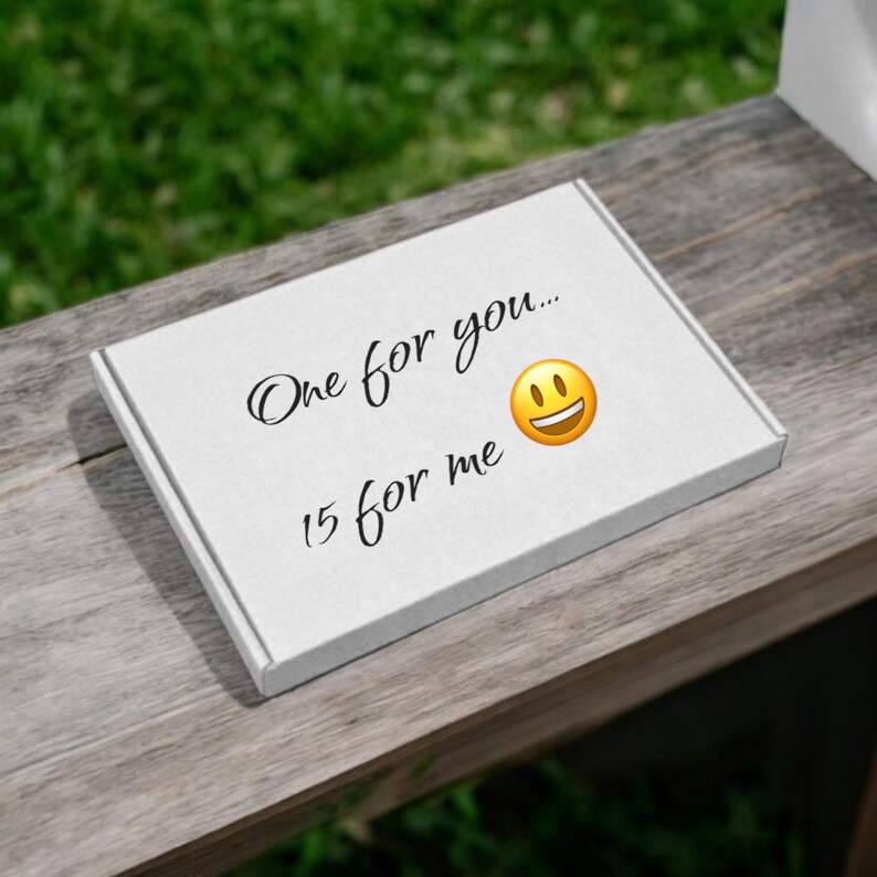 Puede incluir: Una caja rectangular blanca con el texto "One for you..." y "15 for me" en escritura negra. Un emoji de cara sonriente amarilla est&aacute; al lado de las palabras "One for you...". La caja est&aacute; sobre una superficie de madera con un fondo verde.