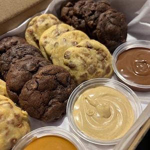 Puede incluir: Una caja de galletas variadas, incluyendo con chispas de chocolate y doble chocolate, con peque&ntilde;os recipientes de salsas para mojar. Las galletas est&aacute;n dispuestas sobre papel blanco dentro de una caja de cart&oacute;n. Las salsas son de chocolate, caramelo y mantequilla de cacahuete.