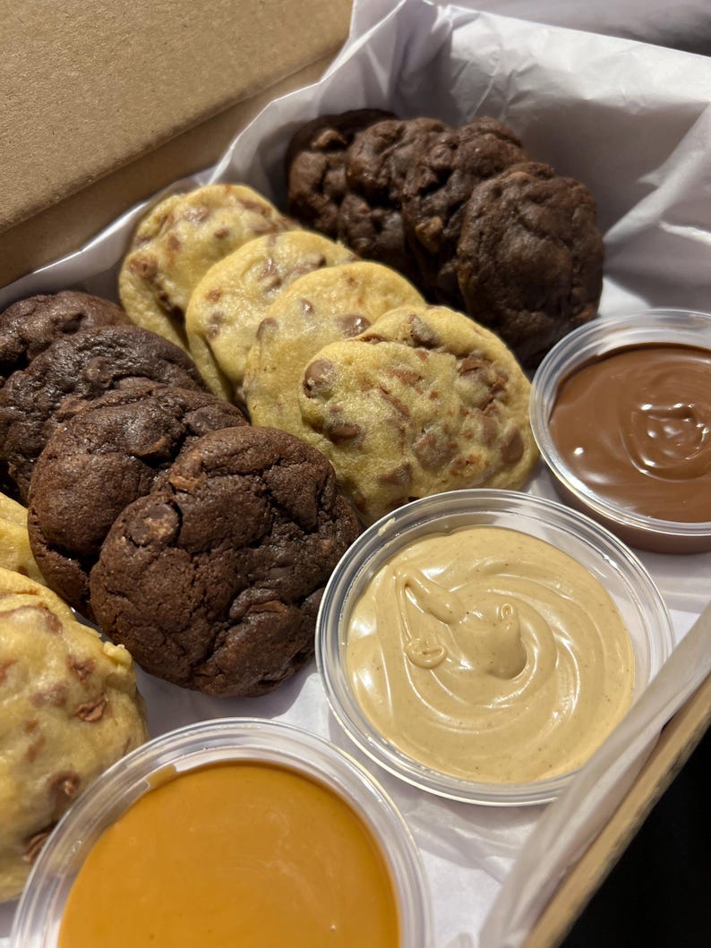 Puede incluir: Una caja de galletas variadas, incluyendo con chispas de chocolate y doble chocolate, con peque&ntilde;os recipientes de salsas para mojar. Las galletas est&aacute;n dispuestas sobre papel blanco dentro de una caja de cart&oacute;n. Las salsas son de chocolate, caramelo y mantequilla de cacahuete.