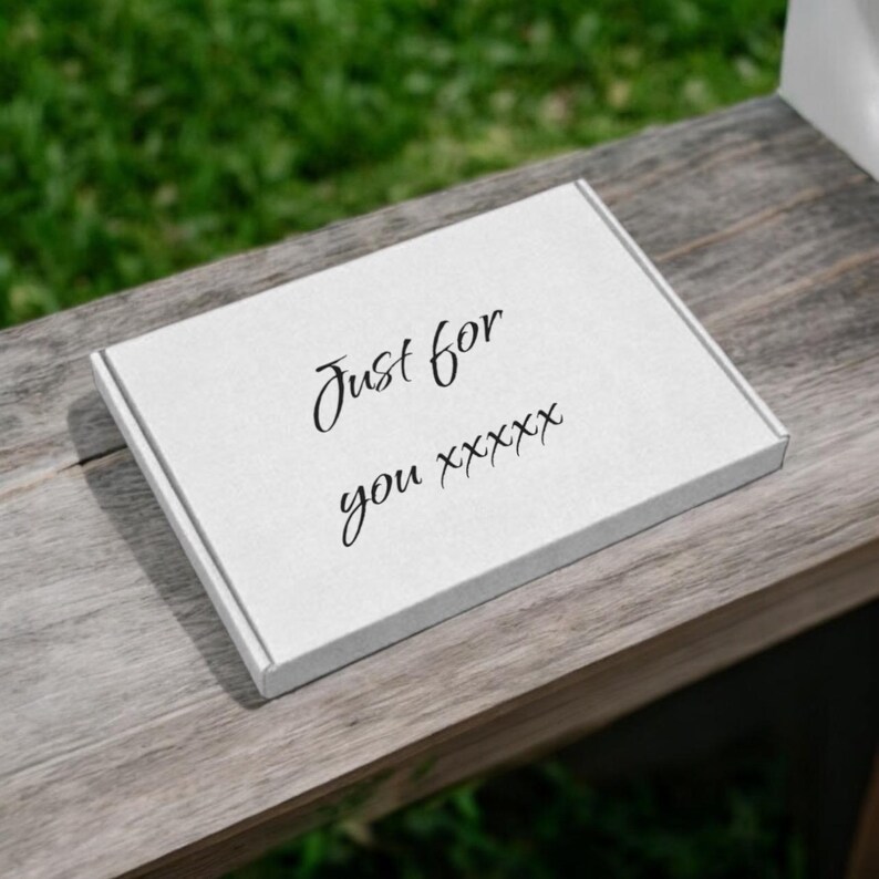 Puede incluir: Una caja rectangular blanca con las palabras "Just for you" en elegante escritura negra. La caja descansa sobre una superficie de madera desgastada, con un fondo verde borroso.