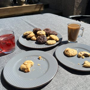 Puede incluir: Una mesa con galletas y bebidas. Varias galletas, algunas de chocolate y otras de vainilla, est&aacute;n en un plato gris. Tambi&eacute;n hay un vaso de l&iacute;quido rojo y un vaso de caf&eacute;. Tambi&eacute;n se ven dos platos adicionales con galletas.