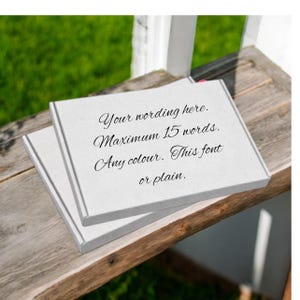 Puede incluir: Dos cajas cuadradas blancas apiladas sobre una superficie de madera envejecida. La caja superior tiene texto en escritura elegante: "Your wording here. Maximum 15 words. Any colour. This font or plain."