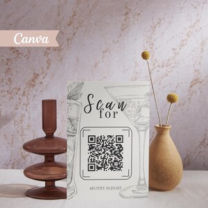 Könnte beinhalten: Eine weiße Karte mit einem QR-Code und dem Text "Scan for Spotify Playlist" wird angezeigt. Die Karte zeigt eine Cocktail-Illustration. Ein brauner Glas-Kerzenhalter und eine kleine Vase mit Trockenblumen sind ebenfalls vorhanden.