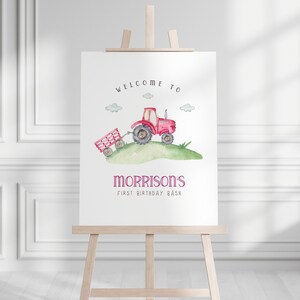 May include: Welcome to Morrison's First Birthday Bash sign featuring a red tractor with a wagon pulling behind it on a green grassy hill. The sign is on an easel.