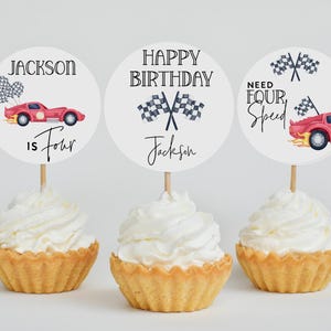 Pu&ograve; includere: Tre cupcake con glassa bianca, ciascuno sormontato da una decorazione di carta rotonda. Le decorazioni presentano grafiche di auto da corsa e il testo "Jackson is Four", "Happy Birthday Jackson" e "Need Four Speed".