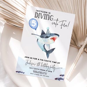 Puede incluir: Una tarjeta de invitación blanca con una ilustración de acuarela de un tiburón con un gorro de fiesta y un globo azul con el número 5. El texto dice "Jackson is Diving into Five!" y los detalles de una fiesta de 5 cumpleaños.