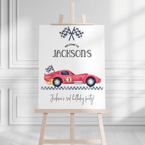 Pu&ograve; includere: Un cartello bianco su un cavalletto di legno presenta un'auto da corsa rossa con il numero 3, bandiere a scacchi e il testo "Welcome to Jackson's" e "Jackson's 3rd birthday party!" in nero.