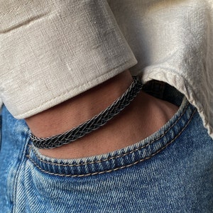 K&ouml;nnte beinhalten: Ein geflochtenes Silberarmband wird am Handgelenk getragen. Das Armband besteht aus miteinander verflochtenen Metallstr&auml;ngen. Die Person tr&auml;gt ein helles Hemd mit Kn&ouml;pfen und eine blaue Jeans. Das Armband ist ein stilvolles Accessoire.