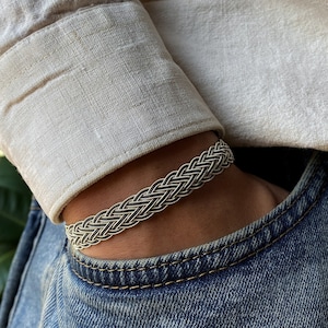 May include: A silver braided bracelet with a woven design. The bracelet is worn on a person's wrist.