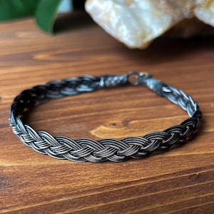 May include: A silver braided bracelet with a dark oxidised finish. The bracelet is made of multiple strands of silver wire that are woven together to create a braided design.