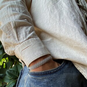 Op de afbeelding: Een zilveren gevlochten armband gedragen op een pols. De armband is gemaakt van meerdere strengen zilverdraad die met elkaar zijn verweven om een gevlochten patroon te cre&euml;ren.