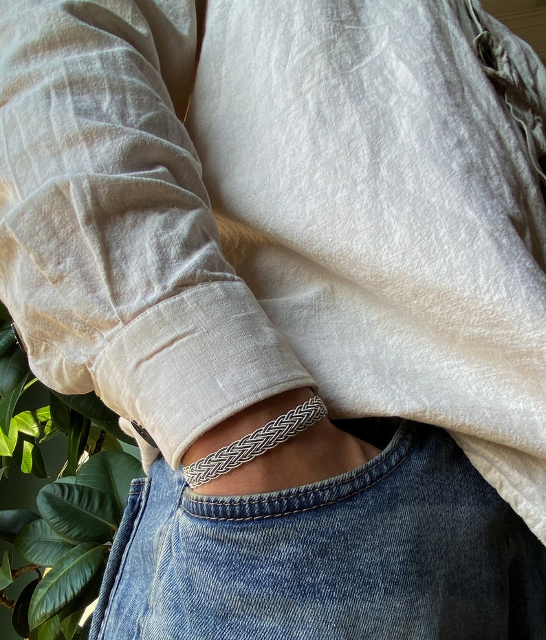 Op de afbeelding: Een zilveren gevlochten armband gedragen op een pols. De armband is gemaakt van meerdere strengen zilverdraad die met elkaar zijn verweven om een gevlochten patroon te cre&euml;ren.