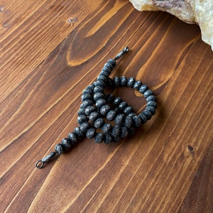 May include: A dark gray beaded necklace with a woven pattern. The beads are round and connected by a dark gray cord. The necklace is lying on a wooden surface.